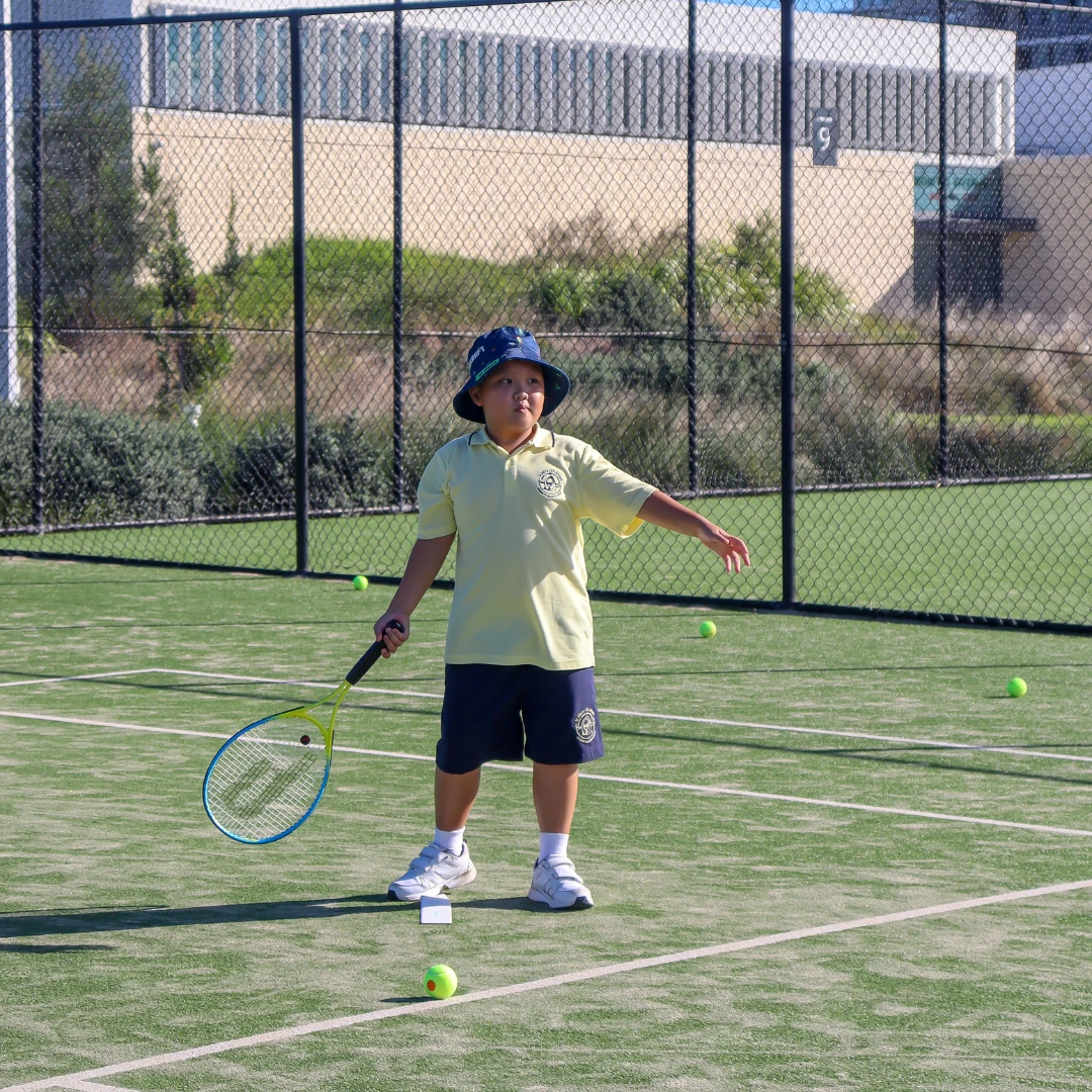 A young child stands on an outdoor tennis court holding a tennis racket after hitting or preparing to hit a tennis ball during a kids' tennis lesson
